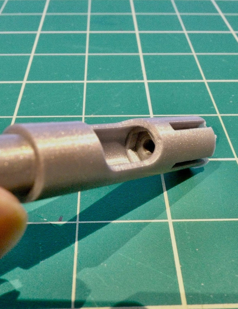 A 3D-printed NoBox stand insert held between finger and thumb on a green cutting mat, showing the printed bore and slot detail