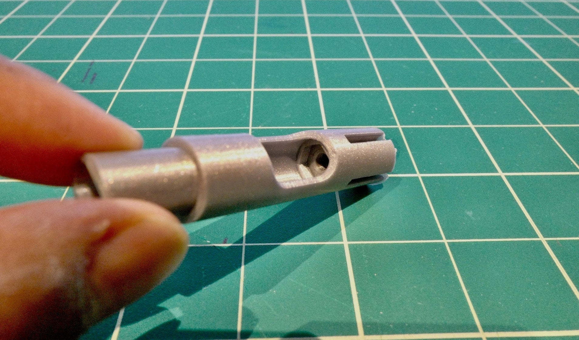 A 3D-printed NoBox stand insert held between finger and thumb on a green cutting mat, showing the printed bore and slot detail