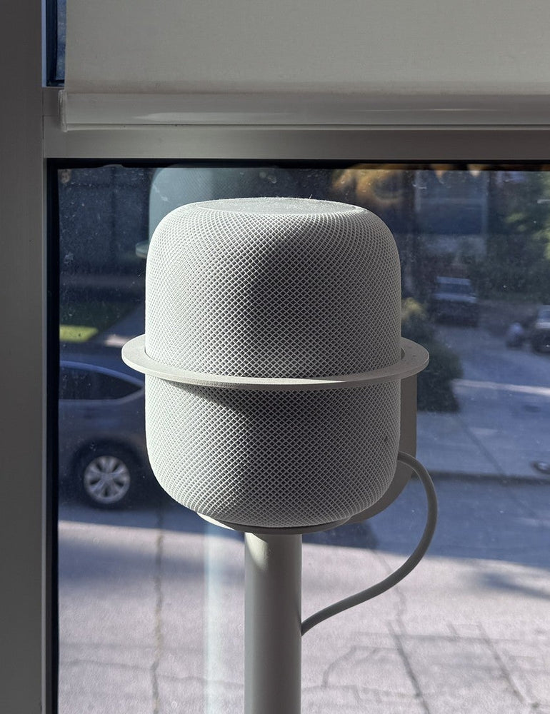 A repaired first-generation HomePod on a NoBox floor stand by a window, its mesh clean, reflected in the glass.