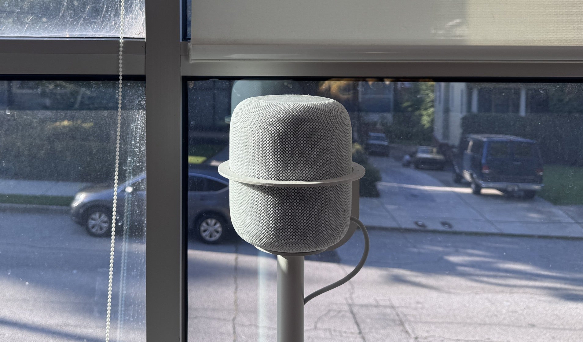 A repaired first-generation HomePod on a NoBox floor stand by a window, its mesh clean, reflected in the glass.