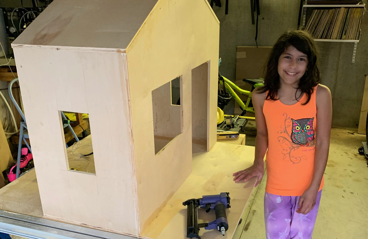 The first NoBox project — a plywood playhouse mid-build on a workshop bench, nail gun beside it, and the young builder who made it